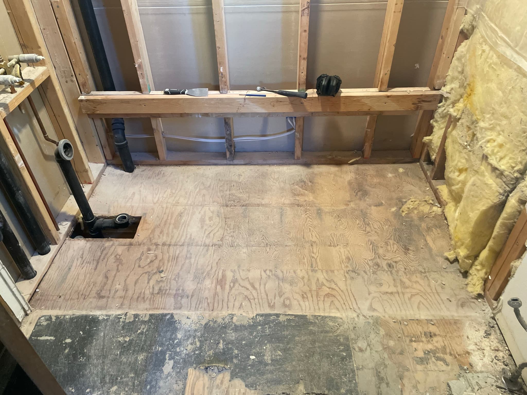 Floor pulled down to plywood subfloor; one wall stripped to studs with insulation and rough plumbing visible.