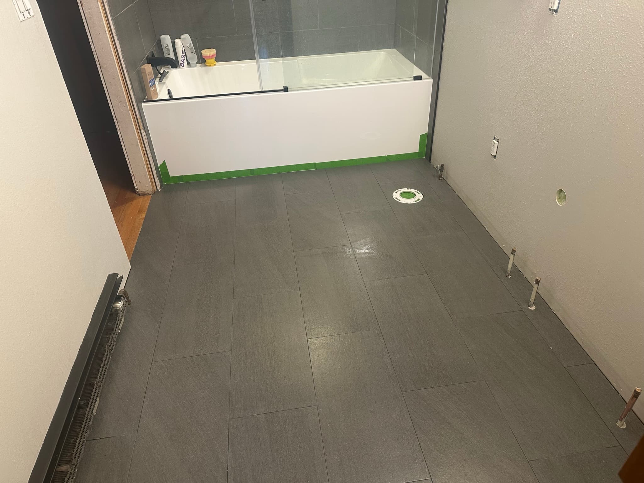 Floor tile laid: dark gray porcelain plank-format running across the room, toilet flange visible center, glass shower door installed on the tub.