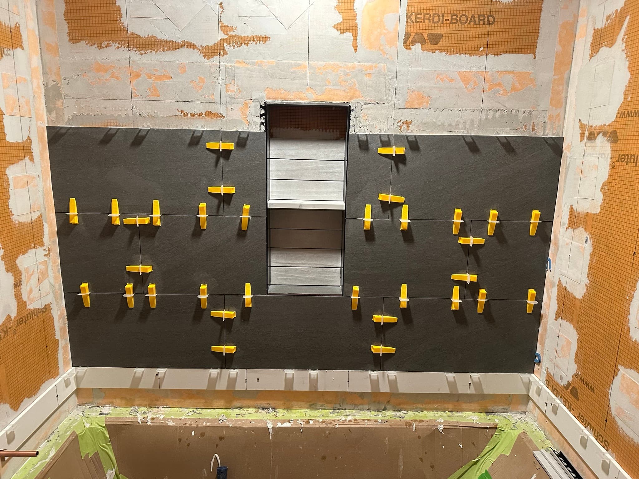 Large-format dark gray porcelain tile going up around the niche with yellow leveling clips on every joint.