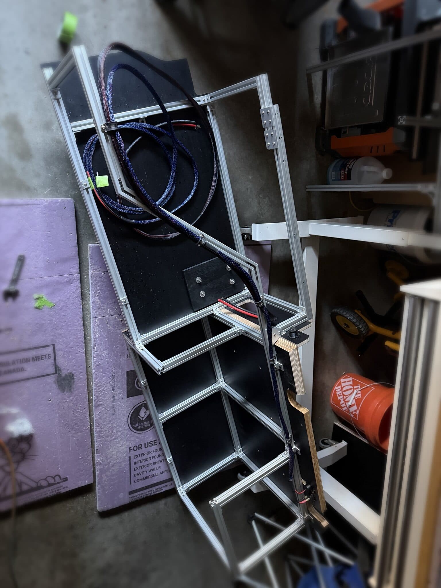 Storage module built from T-slot aluminum extrusion skinned in black laminate plywood, leaning in the garage with braided-sleeve wiring looms routed through the frame.