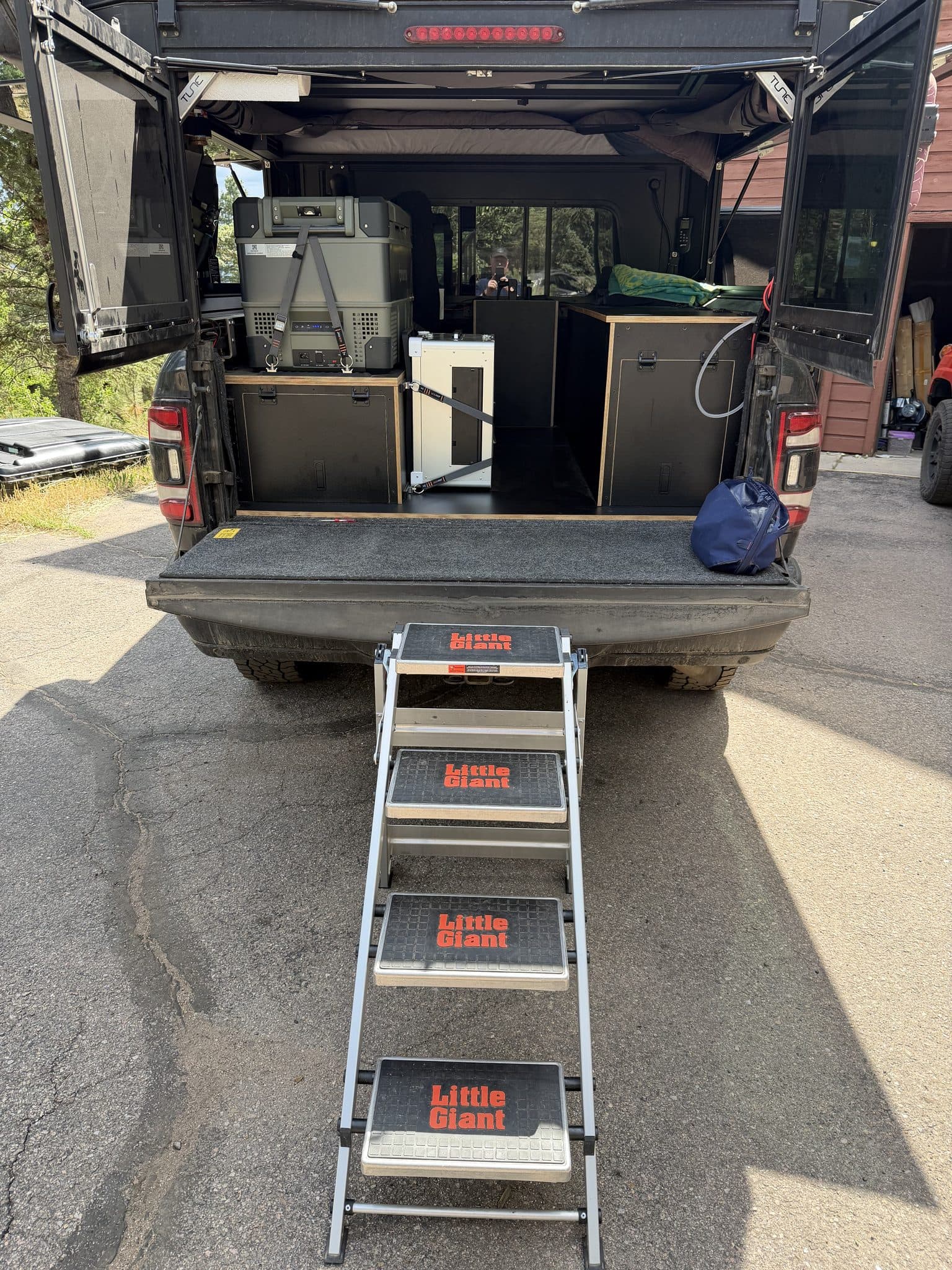 Rear view of the truck with tailgate down and camper doors open: fitted plywood cabinets on both sides, a fridge strapped to the driver-side platform, and folding steps leading up to the bed.