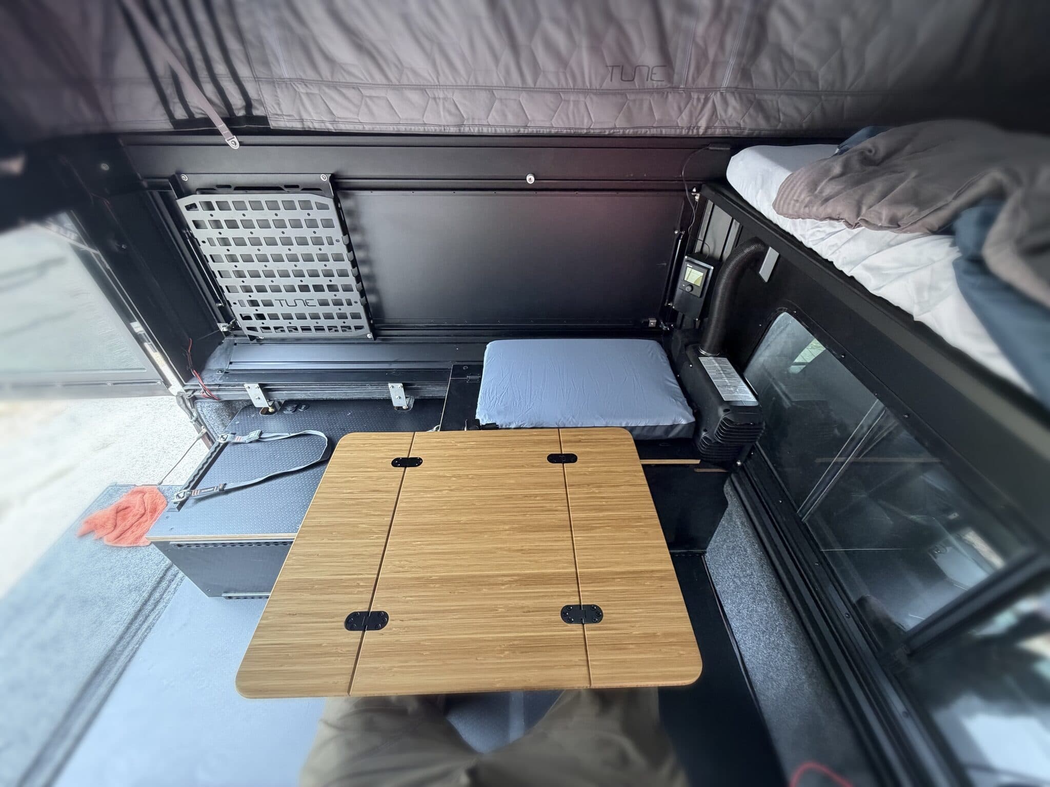 Camper interior with the pop-top up: bamboo Lagun table deployed over the bench, cushion and bunk made up, diesel heater ducting at right.