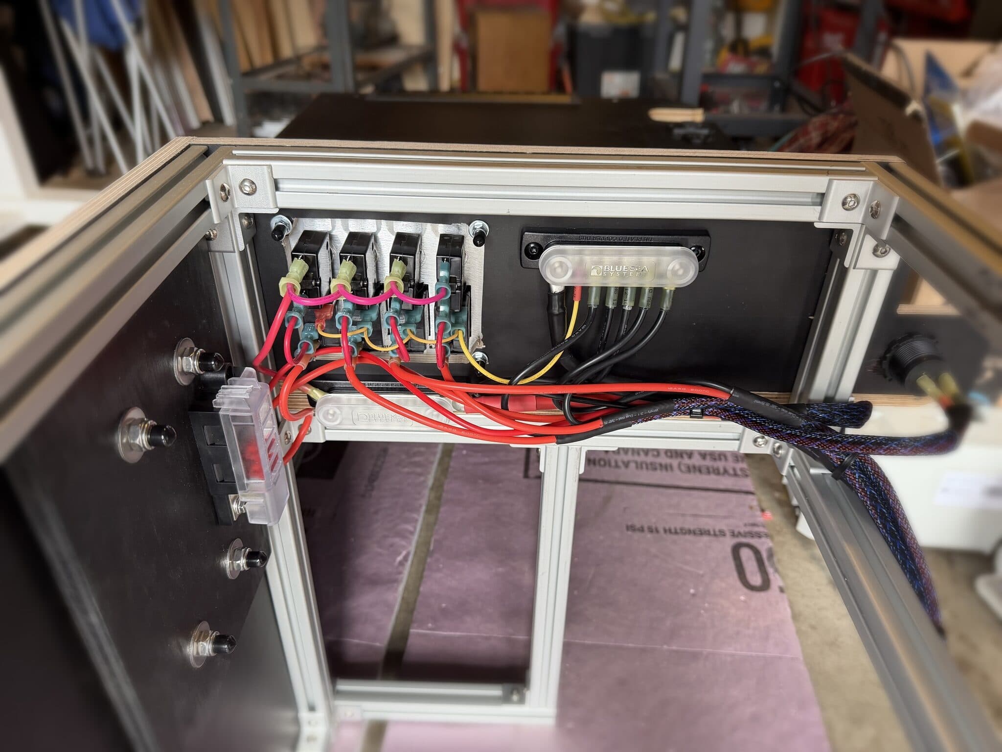Back side of the switch panel: four rocker switches wired with pink and red leads to Blue Sea fuse blocks and a tin-plated busbar, with a 12-volt accessory socket at right.
