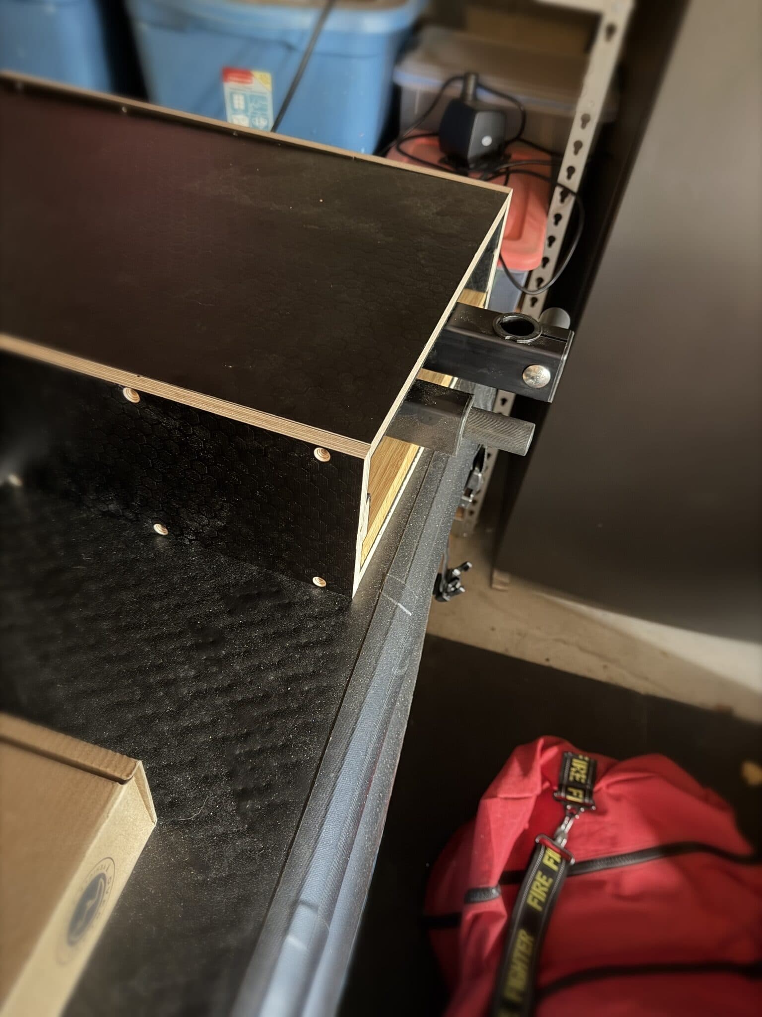 Corner of a hex-laminate plywood bench box with the table arm's square receiver tube clamped to its edge.