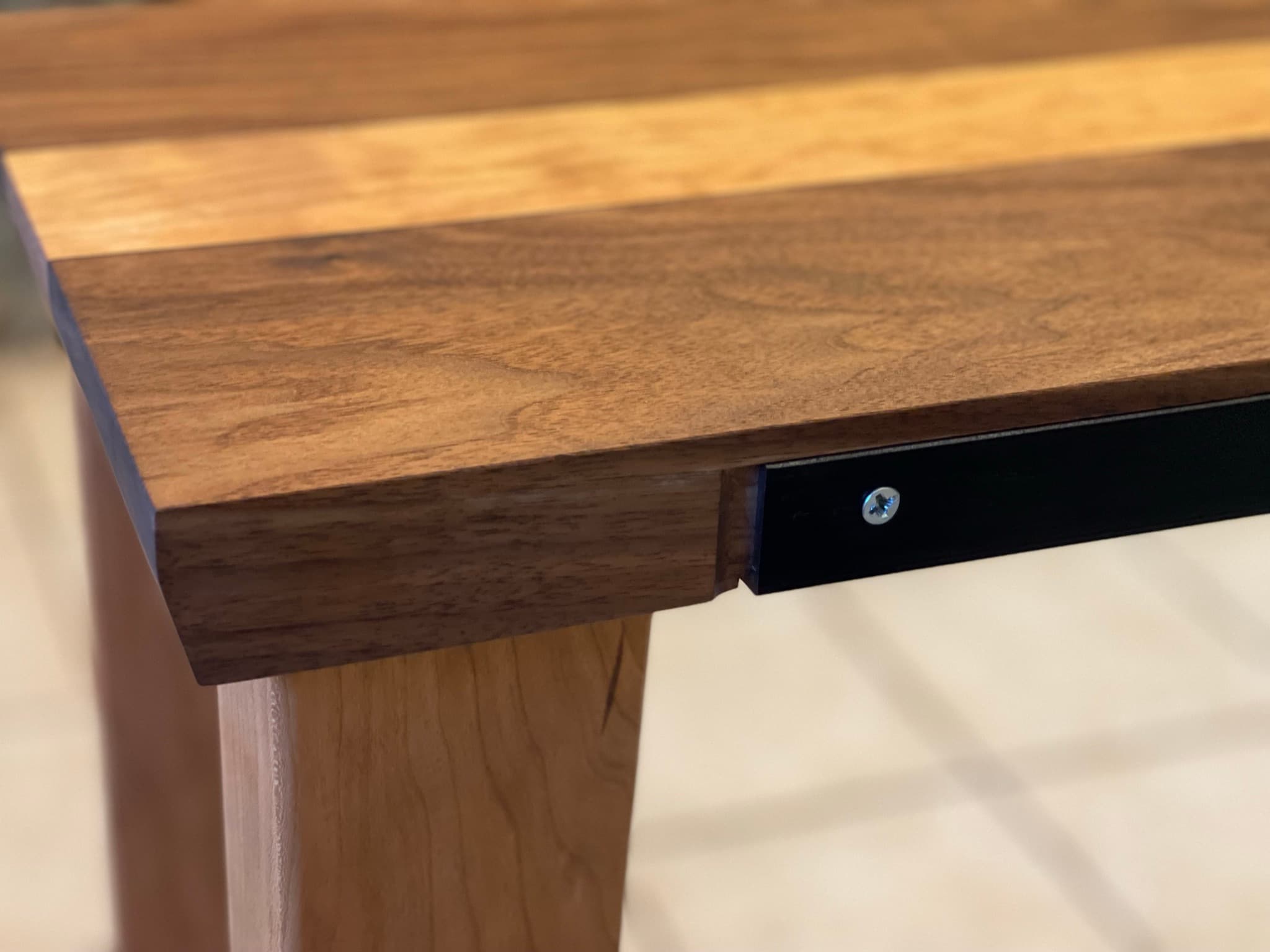 Detail of the rear corner: the blackened-steel back spine inset flush into the top's rear edge, with one tapered maple leg meeting the top.