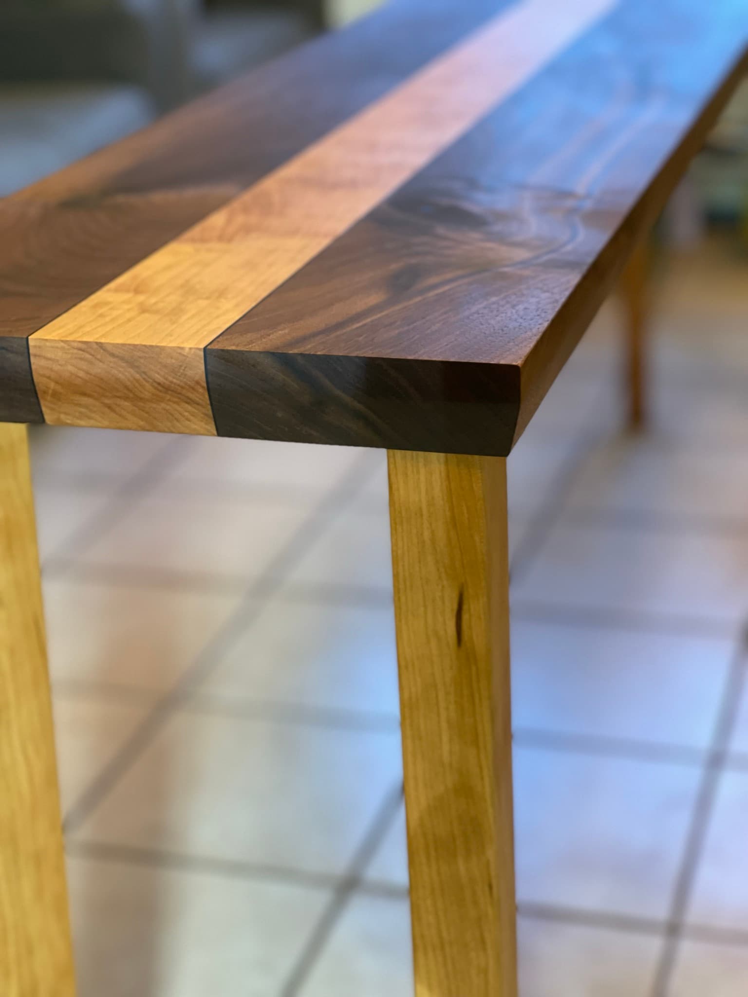 Tighter portrait view of the same corner: chamfered end face, sandwich layers stacked, two maple legs descending to the tile floor.